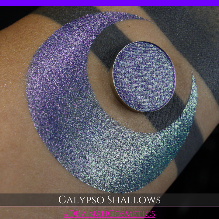 A round eyeshadow pan labeled 'CALYPSO SHALLOWS' made by Devinah Cosmetics, placed over a large crescent moon swatch of the same shimmering shifty eyeshadow on skin. The eyeshadow has a shift between colors gree, purple, blue.