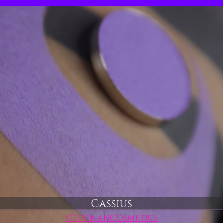 A close-up of a person's arm with a large lavender-purple crescent makeup swatch applied, plus a round pan of matching purple powder resting on the skin. Text at the bottom reads 'CASSIUS' and '#DEVINAHCOSMETICS'.