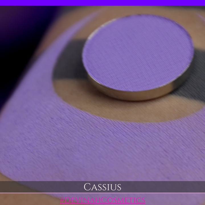 A close-up of a person's arm with a large lavender-purple crescent makeup swatch applied, plus a round pan of matching purple powder resting on the skin. Text at the bottom reads 'CASSIUS' and '#DEVINAHCOSMETICS'.