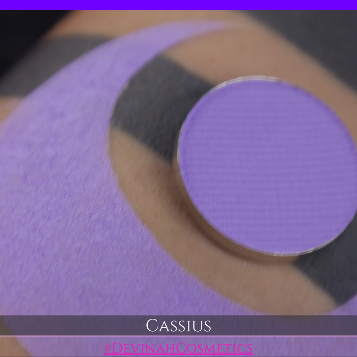 A close-up of a person's arm with a large lavender-purple crescent makeup swatch applied, plus a round pan of matching purple powder resting on the skin. Text at the bottom reads 'CASSIUS' and '#DEVINAHCOSMETICS'.