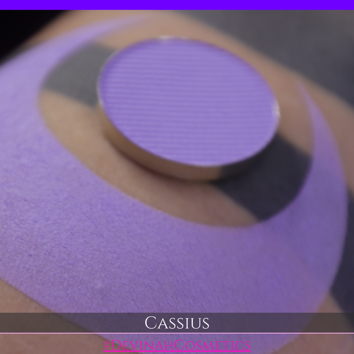 A close-up of a person's arm with a large lavender-purple crescent makeup swatch applied, plus a round pan of matching purple powder resting on the skin. Text at the bottom reads 'CASSIUS' and '#DEVINAHCOSMETICS'.