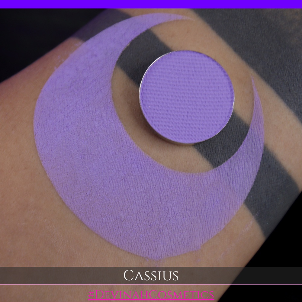 A close-up of a person's arm with a large lavender-purple crescent makeup swatch applied, plus a round pan of matching purple powder resting on the skin. Text at the bottom reads 'CASSIUS' and '#DEVINAHCOSMETICS'.