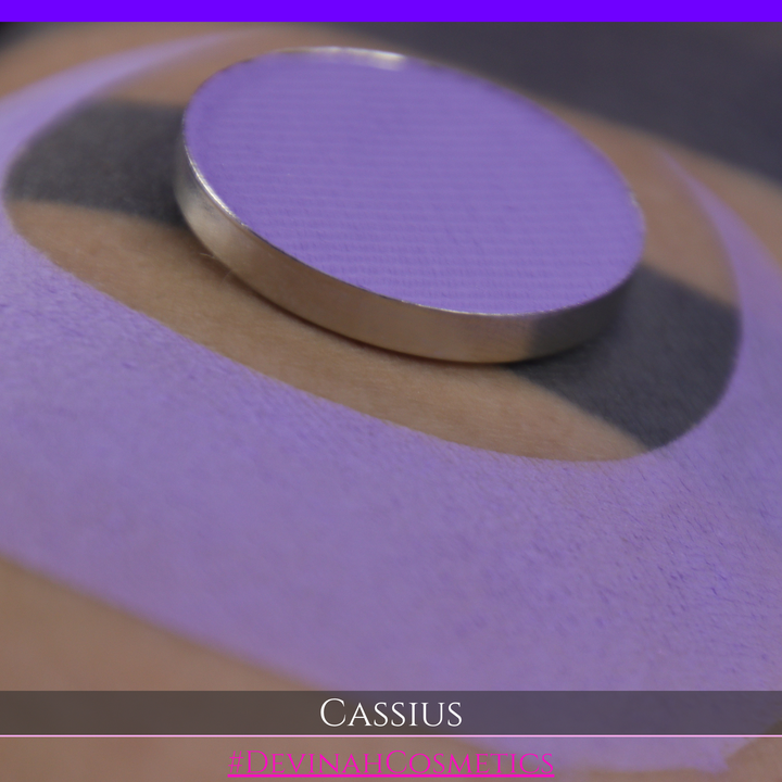 A close-up of a person's arm with a large lavender-purple crescent makeup swatch applied, plus a round pan of matching purple powder resting on the skin. Text at the bottom reads 'CASSIUS' and '#DEVINAHCOSMETICS'.