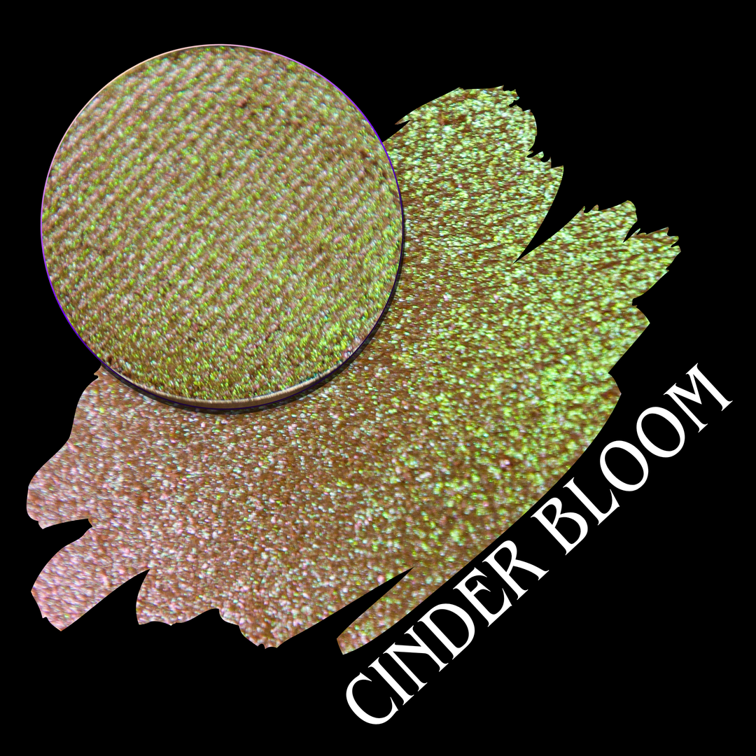 A circular eyeshadow pan and a textured color swatch on a black background, labeled 'CINDER BLOOM' in white text. The shade shifts from warm brown base to bright chartreuse green with pink shimmer made by Devinah Cosmetics.