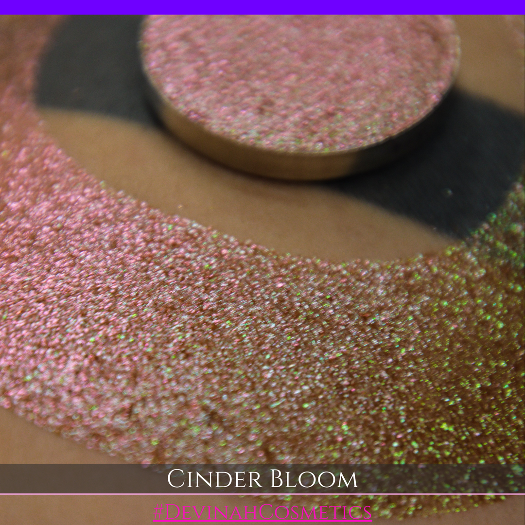 A close-up of a person's arm with a large swatch in a crescent shape and a round eyeshadow pan placed on top. The text overlay reads 'CINDER BLOOM' and '#DEVINAH COSMETICS'.