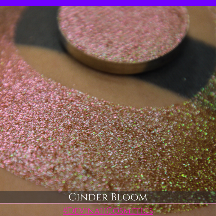 A close-up of a person's arm with a large swatch in a crescent shape and a round eyeshadow pan placed on top. The text overlay reads 'CINDER BLOOM' and '#DEVINAH COSMETICS'.
