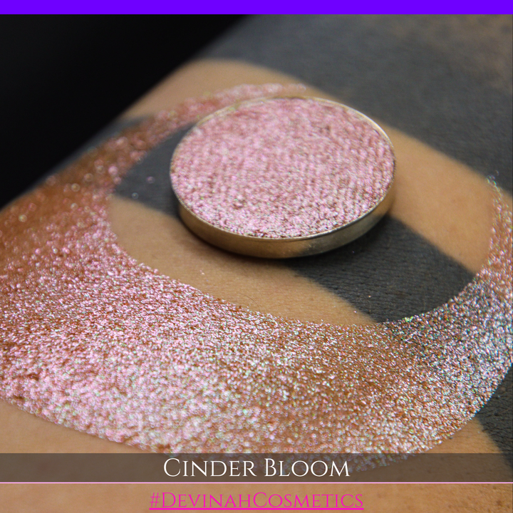 A close-up of a person's arm with a large swatch in a crescent shape and a round eyeshadow pan placed on top. The text overlay reads 'CINDER BLOOM' and '#DEVINAH COSMETICS'.