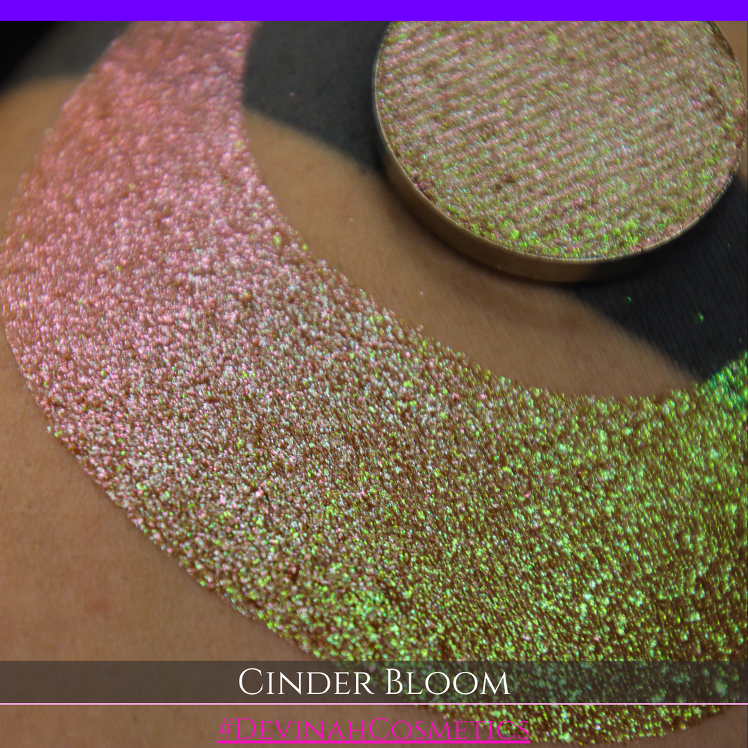 A close-up of a person's arm with a large swatch in a crescent shape and a round eyeshadow pan placed on top. The text overlay reads 'CINDER BLOOM' and '#DEVINAH COSMETICS'.