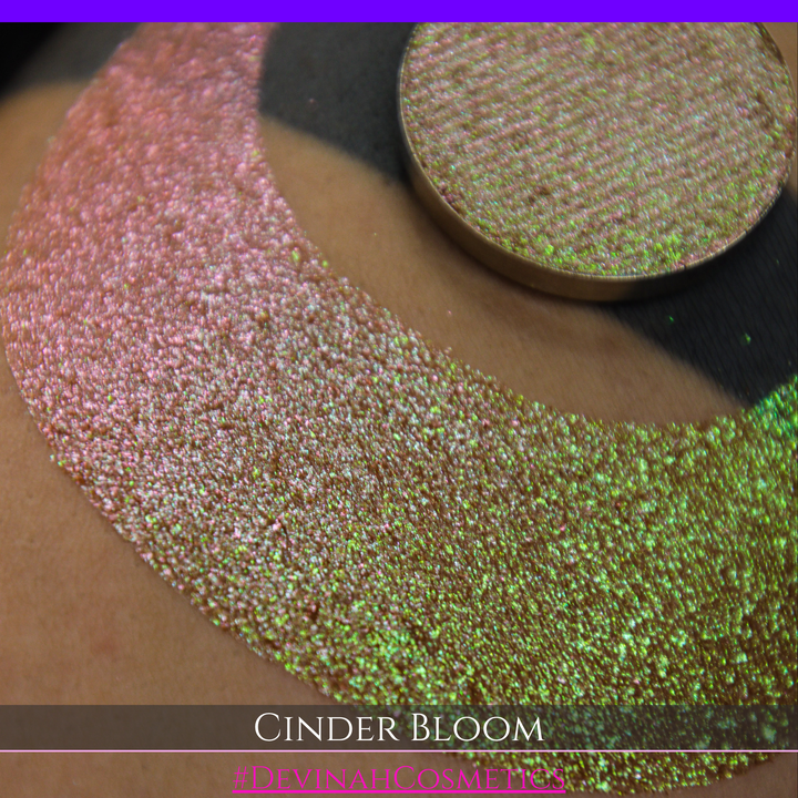 A close-up of a person's arm with a large swatch in a crescent shape and a round eyeshadow pan placed on top. The text overlay reads 'CINDER BLOOM' and '#DEVINAH COSMETICS'.
