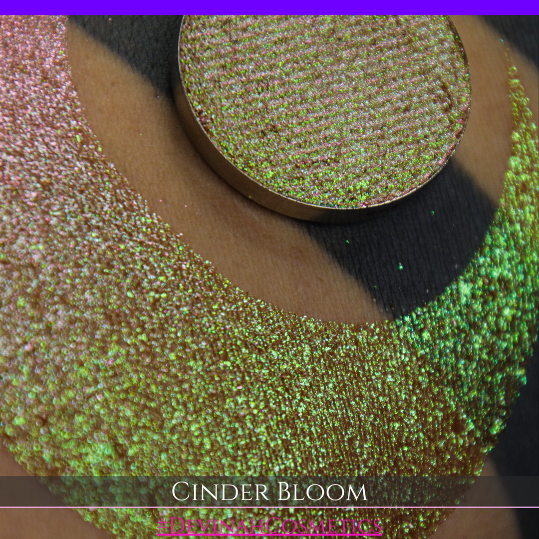 A close-up of a person's arm with a large swatch in a crescent shape and a round eyeshadow pan placed on top. The text overlay reads 'CINDER BLOOM' and '#DEVINAH COSMETICS'.