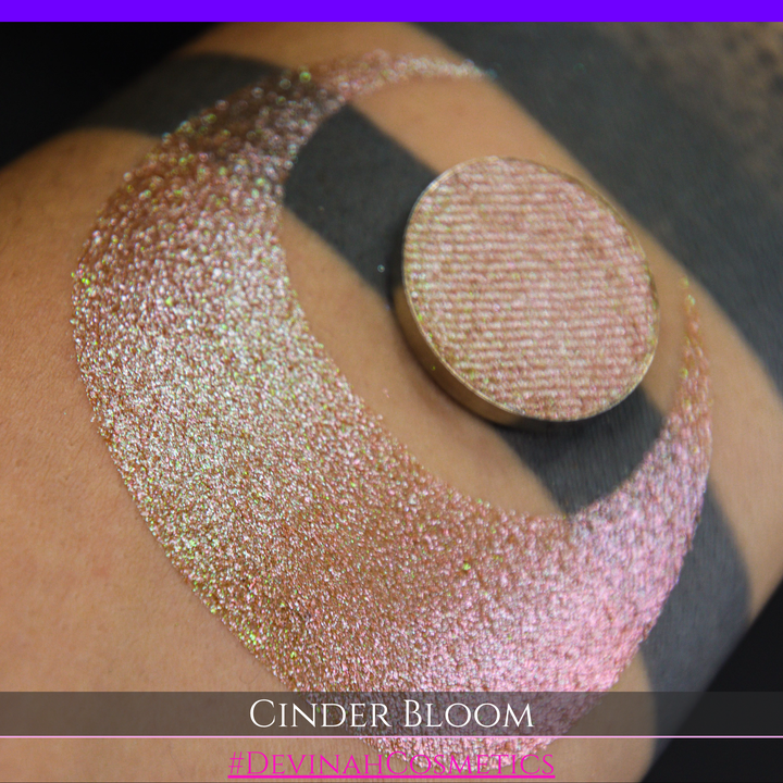 A close-up of a person's arm with a large swatch in a crescent shape and a round eyeshadow pan placed on top. The text overlay reads 'CINDER BLOOM' and '#DEVINAH COSMETICS'.