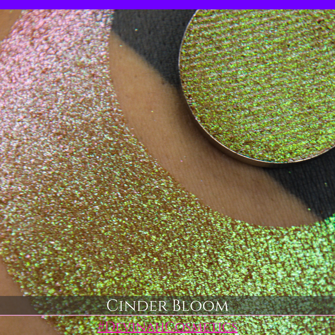 A close-up of a person's arm with a large swatch in a crescent shape and a round eyeshadow pan placed on top. The text overlay reads 'CINDER BLOOM' and '#DEVINAH COSMETICS'.