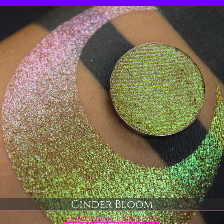 A close-up of a person's arm with a large swatch in a crescent shape and a round eyeshadow pan placed on top. The text overlay reads 'CINDER BLOOM' and '#DEVINAH COSMETICS'.