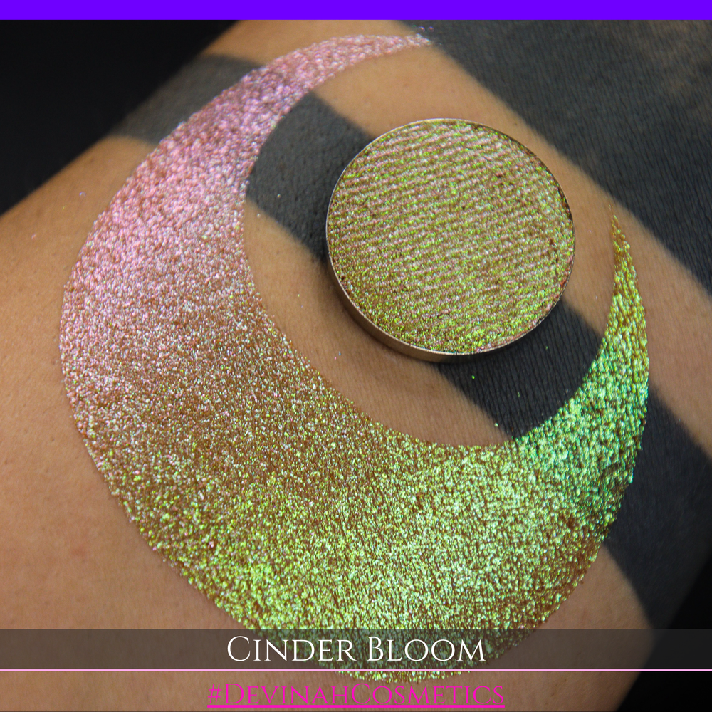 A close-up of a person's arm with a large swatch in a crescent shape and a round eyeshadow pan placed on top. The text overlay reads 'CINDER BLOOM' and '#DEVINAH COSMETICS'.