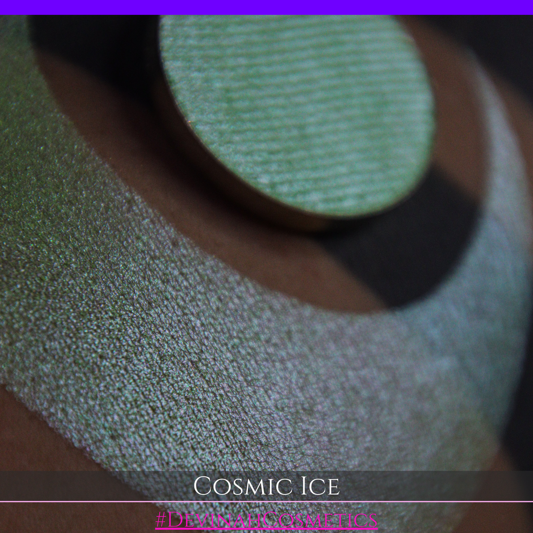 A close-up of a person's arm showing a shimmery green eyeshadow in a circular pan, placed over a larger swatch. Text at the bottom reads 'Cosmic Ice' and '#DevinahCosmetics'.