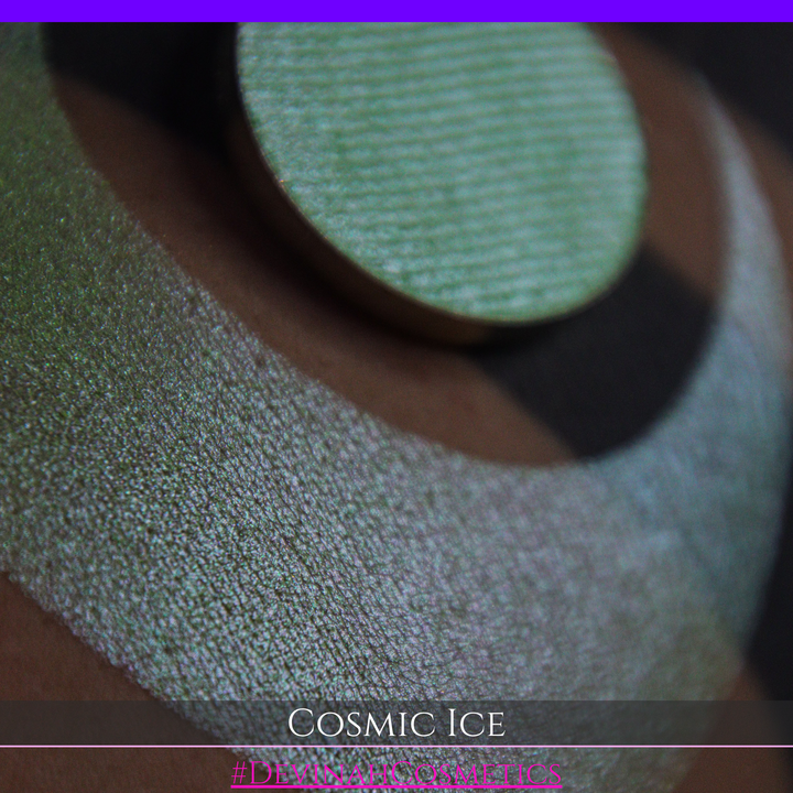 A close-up of a person's arm showing a shimmery green eyeshadow in a circular pan, placed over a larger swatch. Text at the bottom reads 'Cosmic Ice' and '#DevinahCosmetics'.