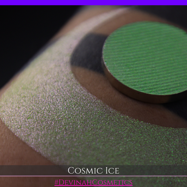 A close-up of a person's arm showing a shimmery green eyeshadow in a circular pan, placed over a larger swatch. Text at the bottom reads 'Cosmic Ice' and '#DevinahCosmetics'.