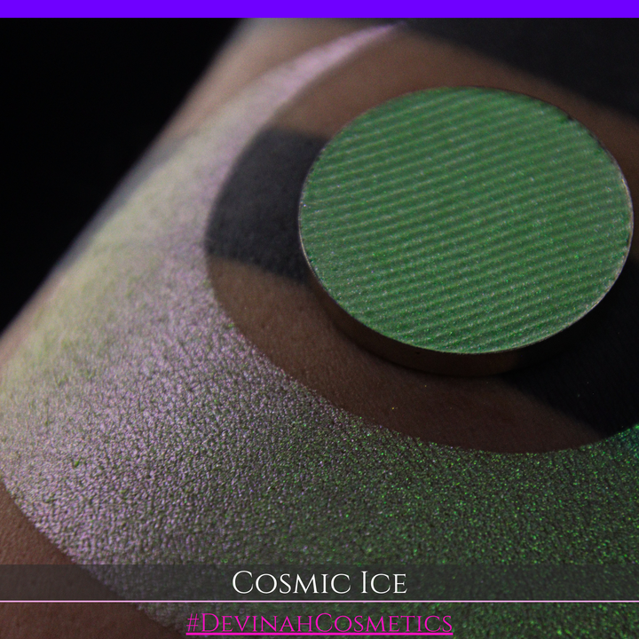 A close-up of a person's arm showing a shimmery green eyeshadow in a circular pan, placed over a larger swatch. Text at the bottom reads 'Cosmic Ice' and '#DevinahCosmetics'.