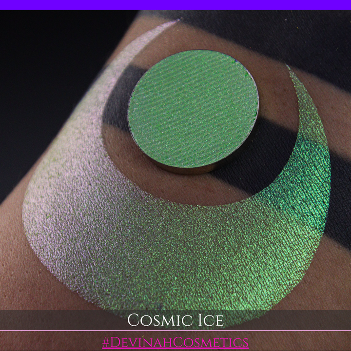 A close-up of a person's arm showing a shimmery green eyeshadow in a circular pan, placed over a larger swatch. Text at the bottom reads 'Cosmic Ice' and '#DevinahCosmetics'.