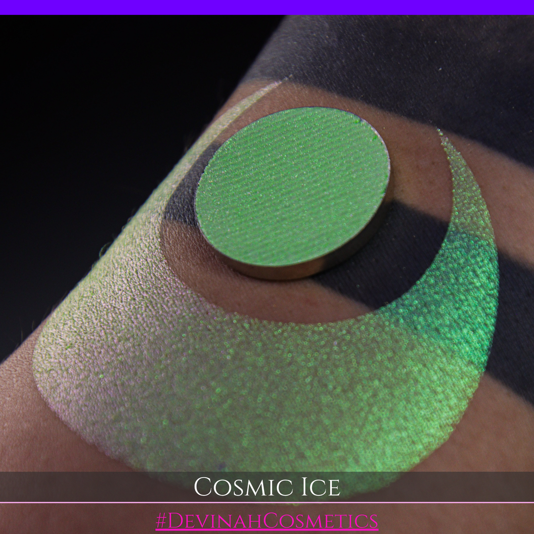 A close-up of a person's arm showing a shimmery green eyeshadow in a circular pan, placed over a larger swatch. Text at the bottom reads 'Cosmic Ice' and '#DevinahCosmetics'.