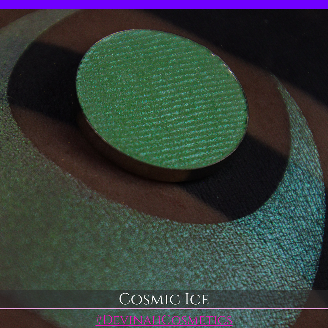 A close-up of a person's arm showing a shimmery green eyeshadow in a circular pan, placed over a larger swatch. Text at the bottom reads 'Cosmic Ice' and '#DevinahCosmetics'.