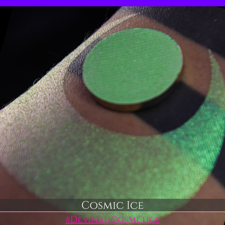A close-up of a person's arm showing a shimmery green eyeshadow in a circular pan, placed over a larger swatch. Text at the bottom reads 'Cosmic Ice' and '#DevinahCosmetics'.