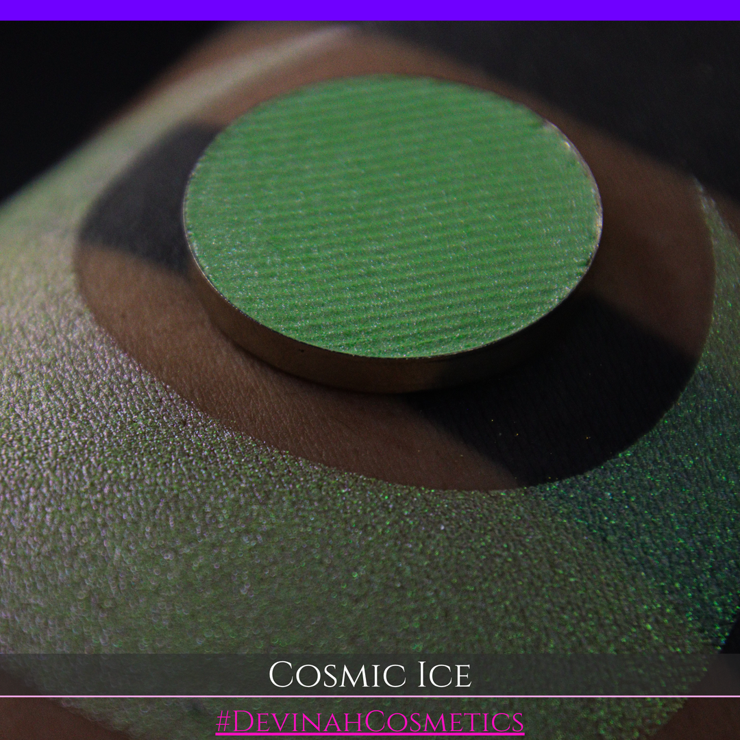 A close-up of a person's arm showing a shimmery green eyeshadow in a circular pan, placed over a larger swatch. Text at the bottom reads 'Cosmic Ice' and '#DevinahCosmetics'.