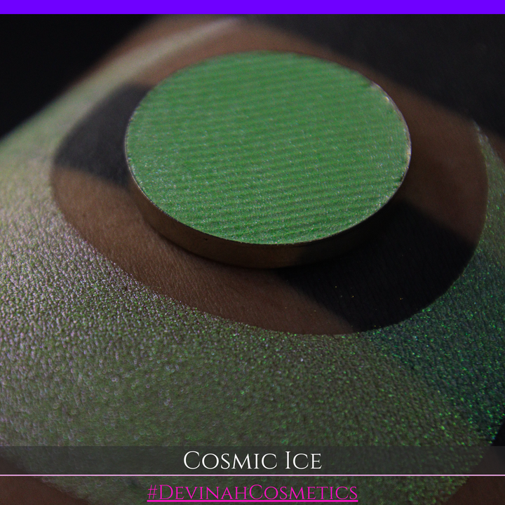 A close-up of a person's arm showing a shimmery green eyeshadow in a circular pan, placed over a larger swatch. Text at the bottom reads 'Cosmic Ice' and '#DevinahCosmetics'.
