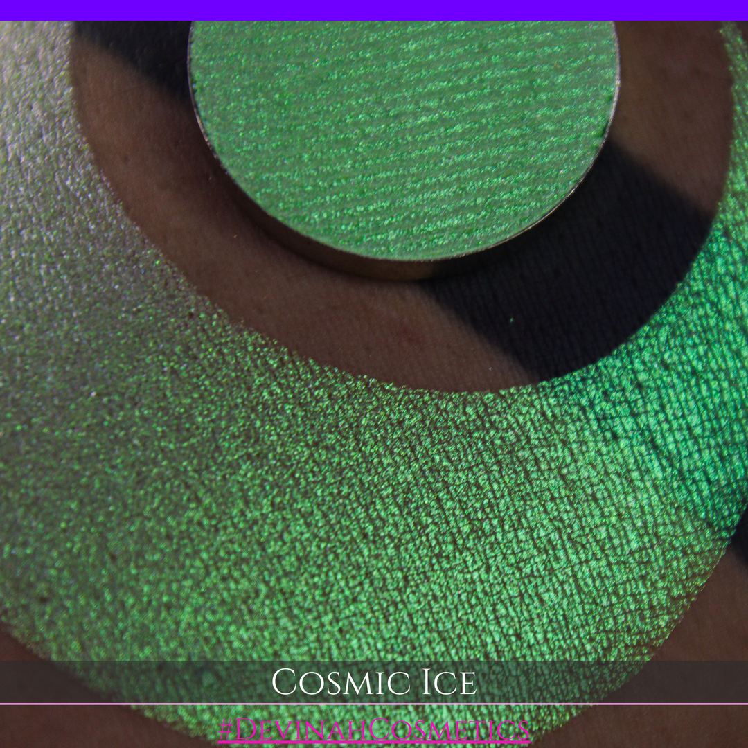 A close-up of a person's arm showing a shimmery green eyeshadow in a circular pan, placed over a larger swatch. Text at the bottom reads 'Cosmic Ice' and '#DevinahCosmetics'.