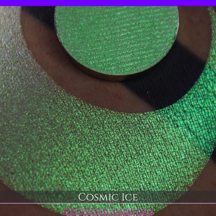 A close-up of a person's arm showing a shimmery green eyeshadow in a circular pan, placed over a larger swatch. Text at the bottom reads 'Cosmic Ice' and '#DevinahCosmetics'.
