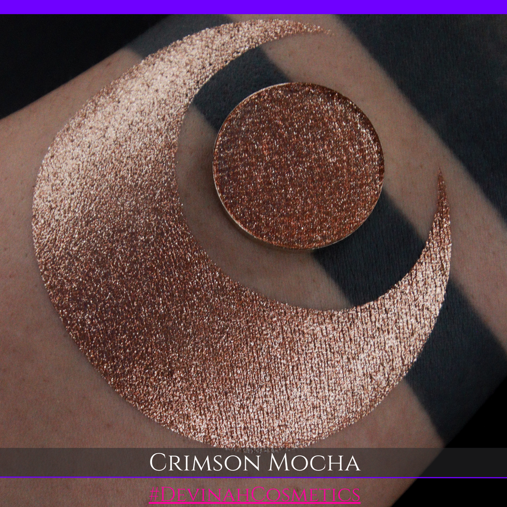 A round eyeshadow pan labeled 'CRIMSON MOCHA' made by Devinah Cosmetics, placed over a large crescent moon swatch of the same shimmering shifty eyeshadow on skin. The eyeshadow has colors of sparkly red and brown shimmer.