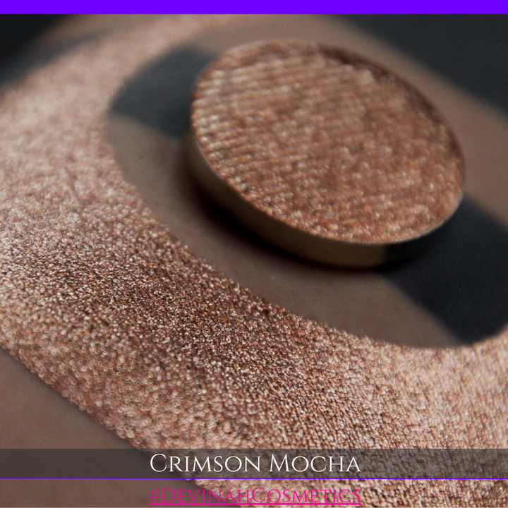 A round eyeshadow pan labeled 'CRIMSON MOCHA' made by Devinah Cosmetics, placed over a large crescent moon swatch of the same shimmering shifty eyeshadow on skin. The eyeshadow has colors of sparkly red and brown shimmer.
