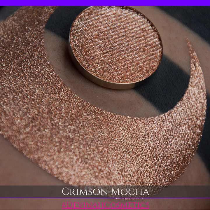 A round eyeshadow pan labeled 'CRIMSON MOCHA' made by Devinah Cosmetics, placed over a large crescent moon swatch of the same shimmering shifty eyeshadow on skin. The eyeshadow has colors of sparkly red and brown shimmer.