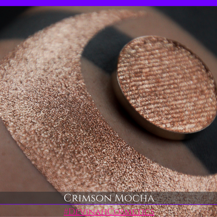 A round eyeshadow pan labeled 'CRIMSON MOCHA' made by Devinah Cosmetics, placed over a large crescent moon swatch of the same shimmering shifty eyeshadow on skin. The eyeshadow has colors of sparkly red and brown shimmer.