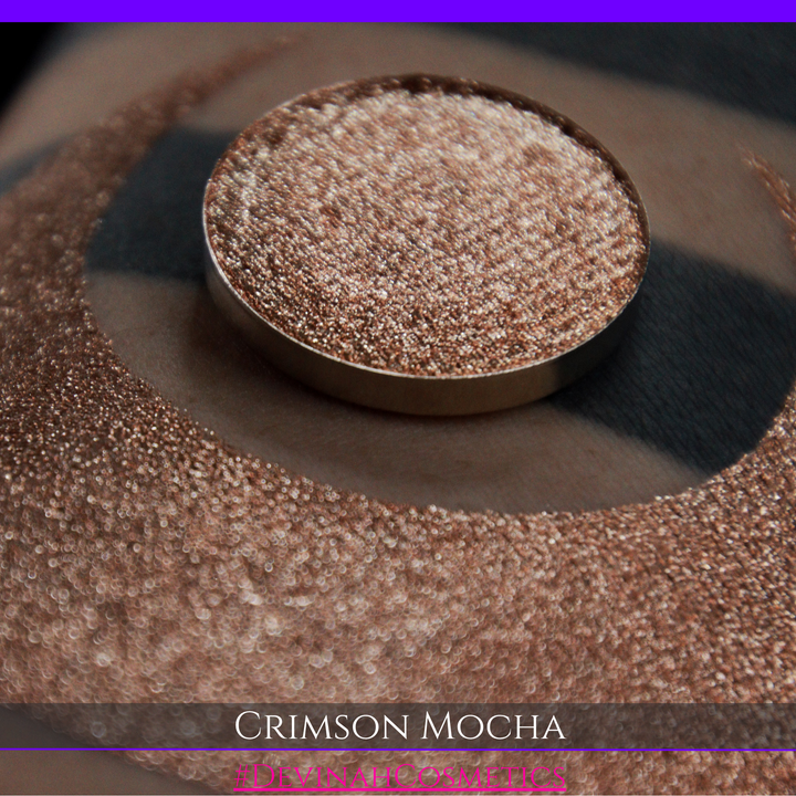 A round eyeshadow pan labeled 'CRIMSON MOCHA' made by Devinah Cosmetics, placed over a large crescent moon swatch of the same shimmering shifty eyeshadow on skin. The eyeshadow has colors of sparkly red and brown shimmer.