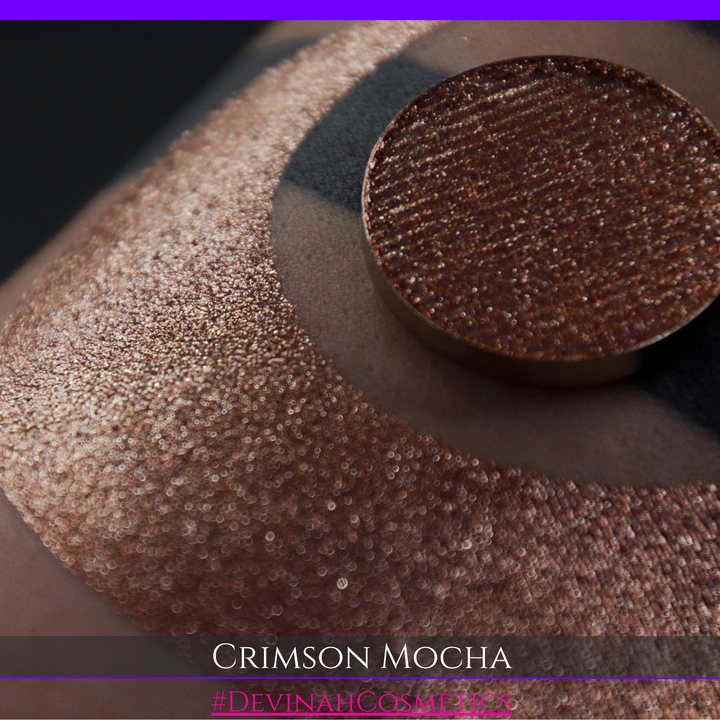 A round eyeshadow pan labeled 'CRIMSON MOCHA' made by Devinah Cosmetics, placed over a large crescent moon swatch of the same shimmering shifty eyeshadow on skin. The eyeshadow has colors of sparkly red and brown shimmer.