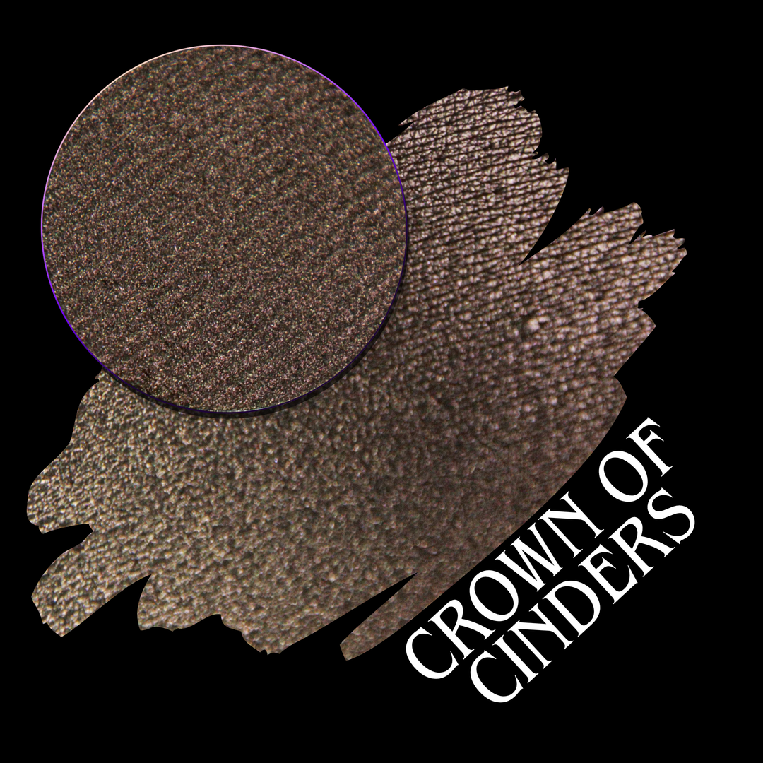 A round eyeshadow pan labeled 'CROWN OF CINDERS' made by Devinah Cosmetics, placed over a large crescent moon swatch of the same shimmering shifty eyeshadow on skin. The eyeshadow has a duochrome multichrome shift between colors bronze, pink, red.