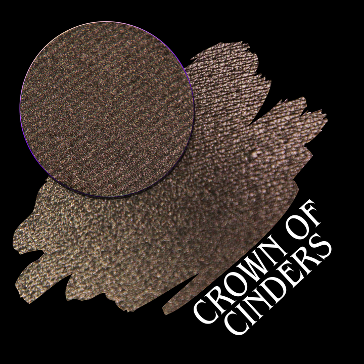 A round eyeshadow pan labeled 'CROWN OF CINDERS' made by Devinah Cosmetics, placed over a large crescent moon swatch of the same shimmering shifty eyeshadow on skin. The eyeshadow has a duochrome multichrome shift between colors bronze, pink, red.