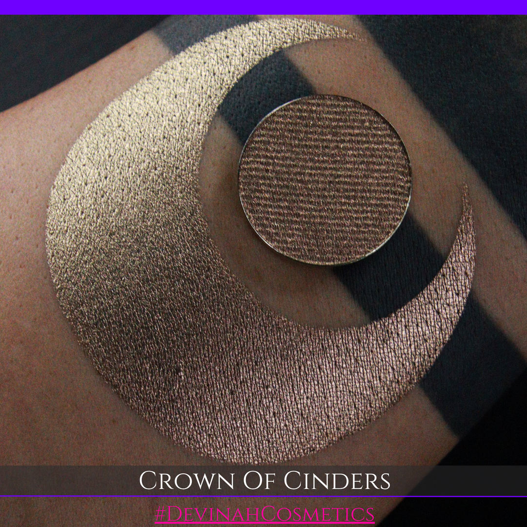 A round eyeshadow pan labeled 'CROWN OF CINDERS' made by Devinah Cosmetics, placed over a large crescent moon swatch of the same shimmering shifty eyeshadow on skin. The eyeshadow has a duochrome multichrome shift between colors bronze, pink, red.