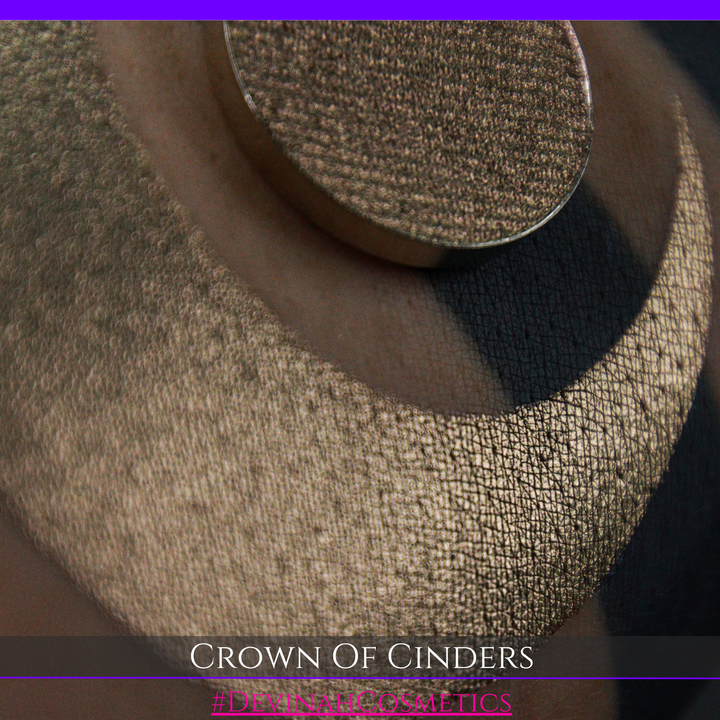 A round eyeshadow pan labeled 'CROWN OF CINDERS' made by Devinah Cosmetics, placed over a large crescent moon swatch of the same shimmering shifty eyeshadow on skin. The eyeshadow has a duochrome multichrome shift between colors bronze, pink, red.