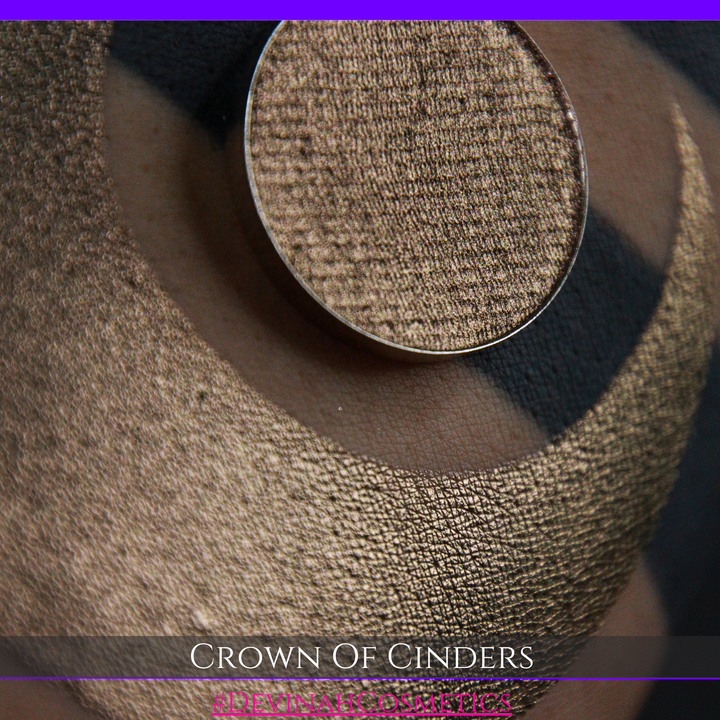 A round eyeshadow pan labeled 'CROWN OF CINDERS' made by Devinah Cosmetics, placed over a large crescent moon swatch of the same shimmering shifty eyeshadow on skin. The eyeshadow has a duochrome multichrome shift between colors bronze, pink, red.