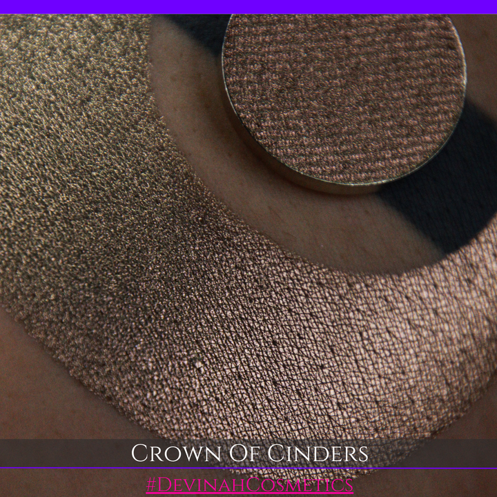 A round eyeshadow pan labeled 'CROWN OF CINDERS' made by Devinah Cosmetics, placed over a large crescent moon swatch of the same shimmering shifty eyeshadow on skin. The eyeshadow has a duochrome multichrome shift between colors bronze, pink, red.