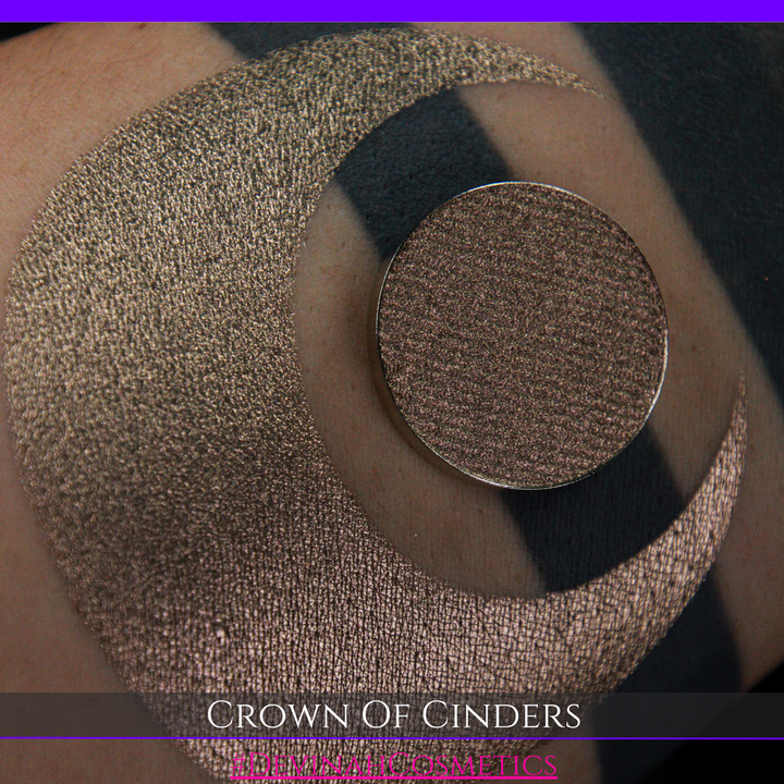 A round eyeshadow pan labeled 'CROWN OF CINDERS' made by Devinah Cosmetics, placed over a large crescent moon swatch of the same shimmering shifty eyeshadow on skin. The eyeshadow has a duochrome multichrome shift between colors bronze, pink, red.
