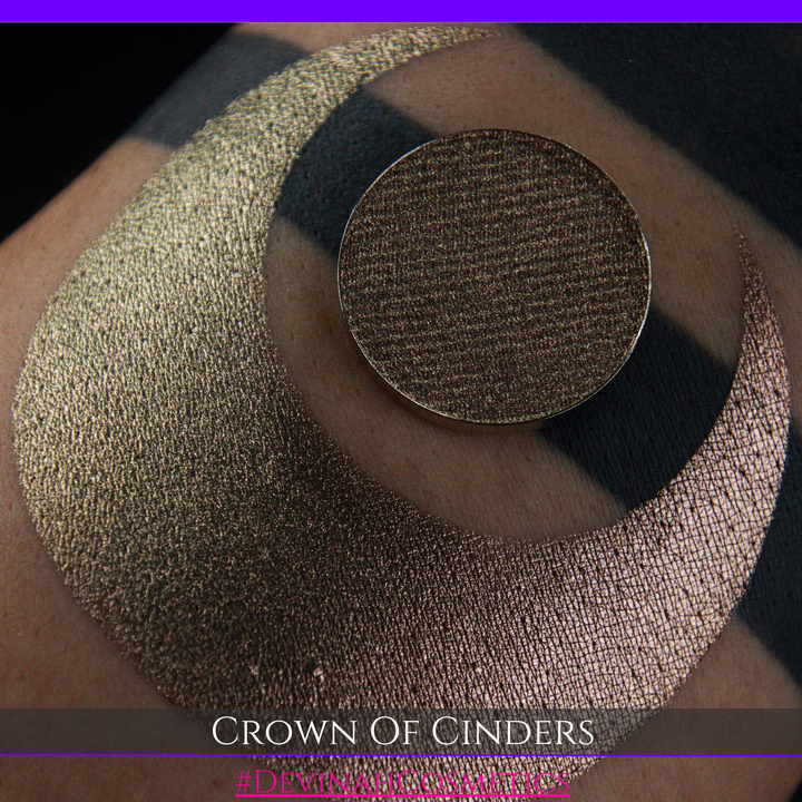 A round eyeshadow pan labeled 'CROWN OF CINDERS' made by Devinah Cosmetics, placed over a large crescent moon swatch of the same shimmering shifty eyeshadow on skin. The eyeshadow has a duochrome multichrome shift between colors bronze, pink, red.