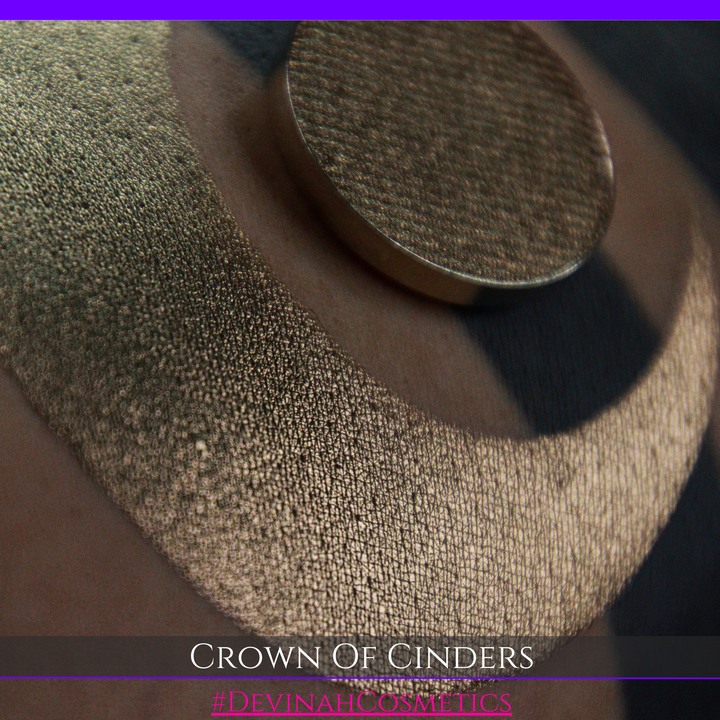 A round eyeshadow pan labeled 'CROWN OF CINDERS' made by Devinah Cosmetics, placed over a large crescent moon swatch of the same shimmering shifty eyeshadow on skin. The eyeshadow has a duochrome multichrome shift between colors bronze, pink, red.