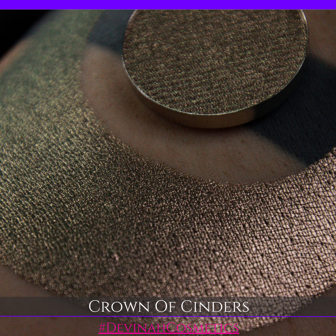 A round eyeshadow pan labeled 'CROWN OF CINDERS' made by Devinah Cosmetics, placed over a large crescent moon swatch of the same shimmering shifty eyeshadow on skin. The eyeshadow has a duochrome multichrome shift between colors bronze, pink, red.
