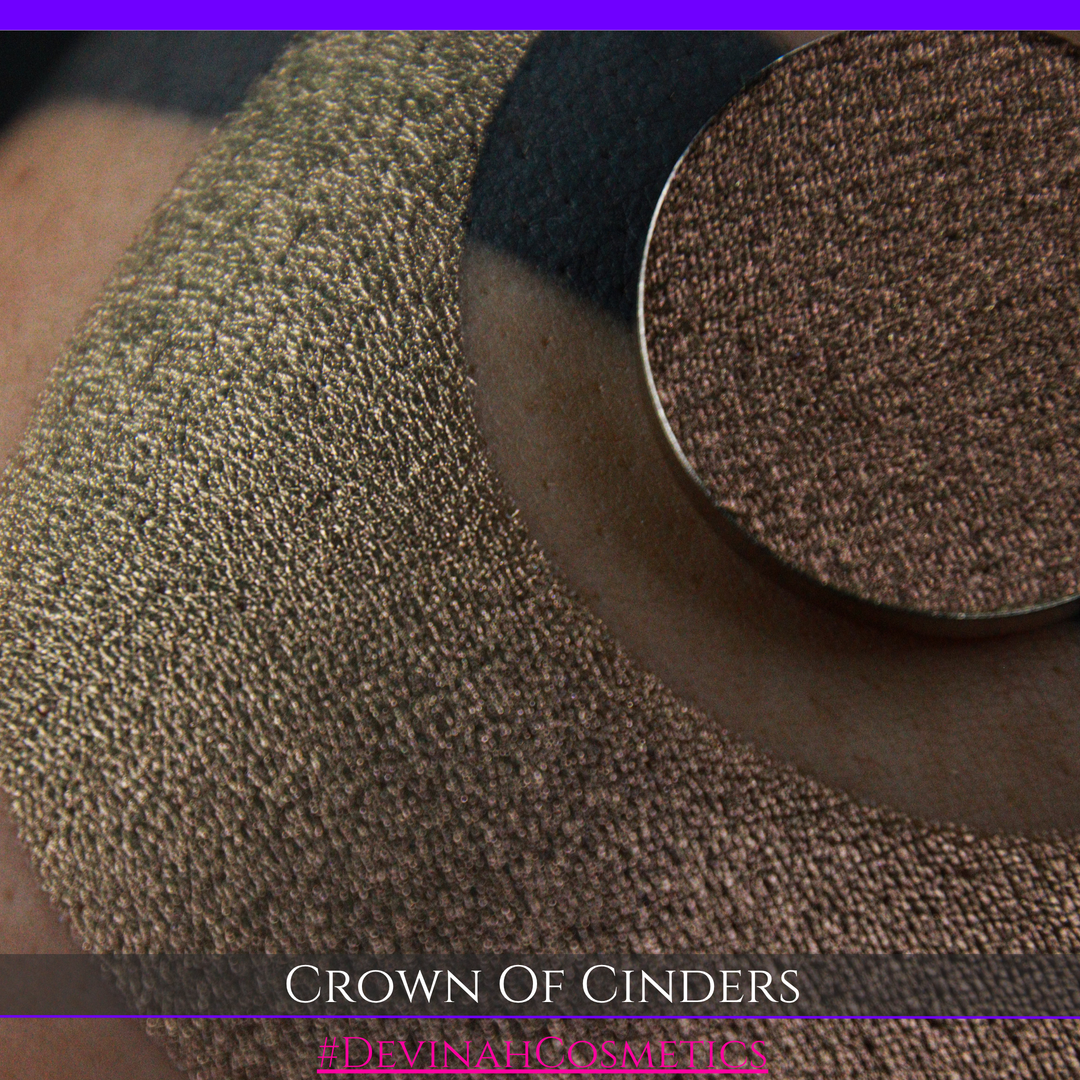 A round eyeshadow pan labeled 'CROWN OF CINDERS' made by Devinah Cosmetics, placed over a large crescent moon swatch of the same shimmering shifty eyeshadow on skin. The eyeshadow has a duochrome multichrome shift between colors bronze, pink, red.