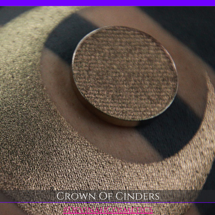 A round eyeshadow pan labeled 'CROWN OF CINDERS' made by Devinah Cosmetics, placed over a large crescent moon swatch of the same shimmering shifty eyeshadow on skin. The eyeshadow has a duochrome multichrome shift between colors bronze, pink, red.