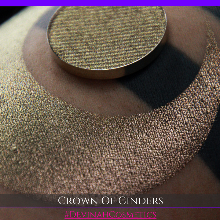 A round eyeshadow pan labeled 'CROWN OF CINDERS' made by Devinah Cosmetics, placed over a large crescent moon swatch of the same shimmering shifty eyeshadow on skin. The eyeshadow has a duochrome multichrome shift between colors bronze, pink, red.