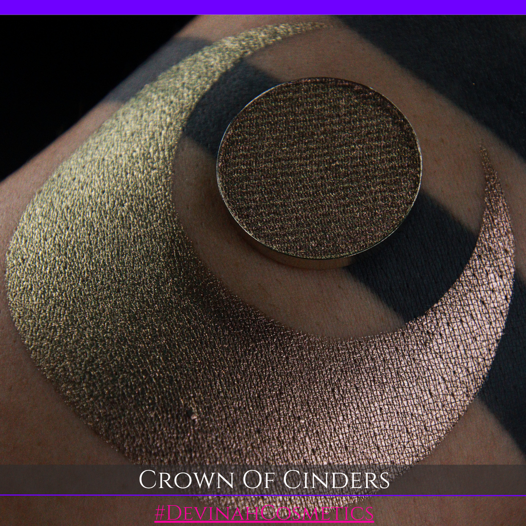 A round eyeshadow pan labeled 'CROWN OF CINDERS' made by Devinah Cosmetics, placed over a large crescent moon swatch of the same shimmering shifty eyeshadow on skin. The eyeshadow has a duochrome multichrome shift between colors bronze, pink, red.